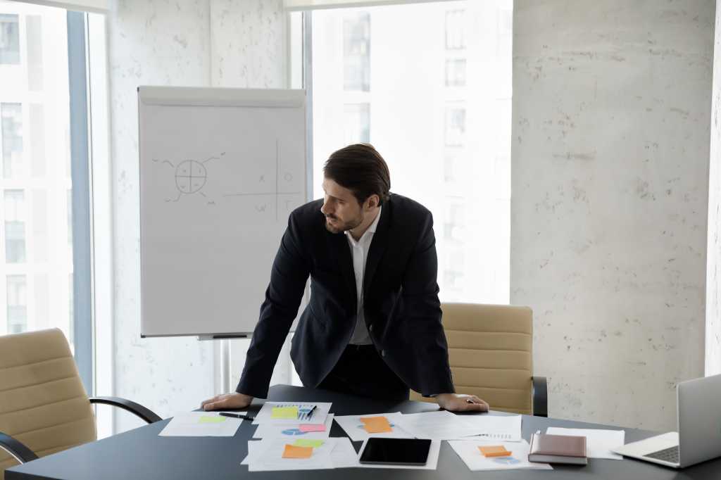 Frustrated concerned business man standing at table with sales reports, paper sticky notes, looking away, thinking over company future, negative statistics result, financial loss